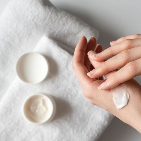 Hands applying white cream beside two open skincare jars on a white towel