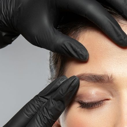 Gloved hands shaping a person’s eyebrow with eyes closed during a beauty treatment