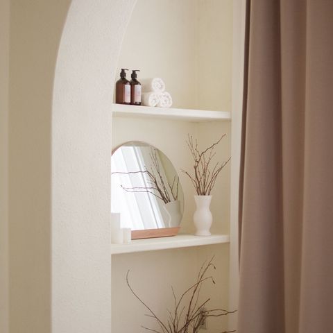 Minimal beige shelves with bottles, a mirror, and dried branches in white vases on a wall niche
