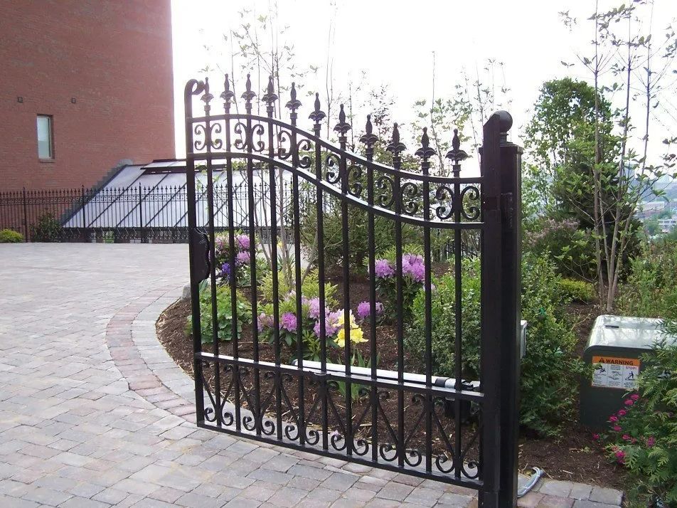 Black wrought iron gate partially open to a garden with brick pathway.