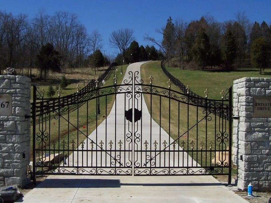 Black wrought-iron gate at the entrance to a long, paved driveway, flanked by stone pillars and grassy hills.