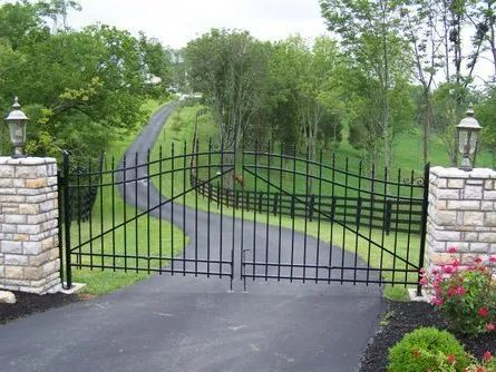 Black wrought-iron gate between stone pillars, opening to a paved driveway. Green trees in the background.
