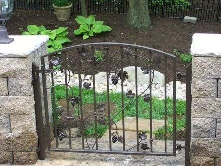 Wrought iron garden gate with grape vine design, set between stone pillars. Green foliage in background.