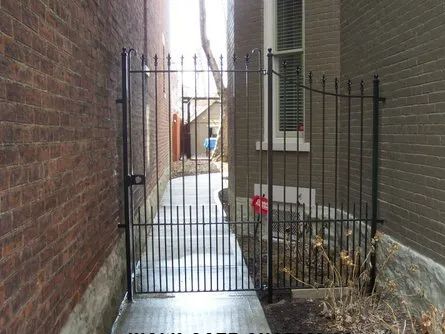 Black metal gate in a narrow alleyway between two brick buildings, leading to a walkway.