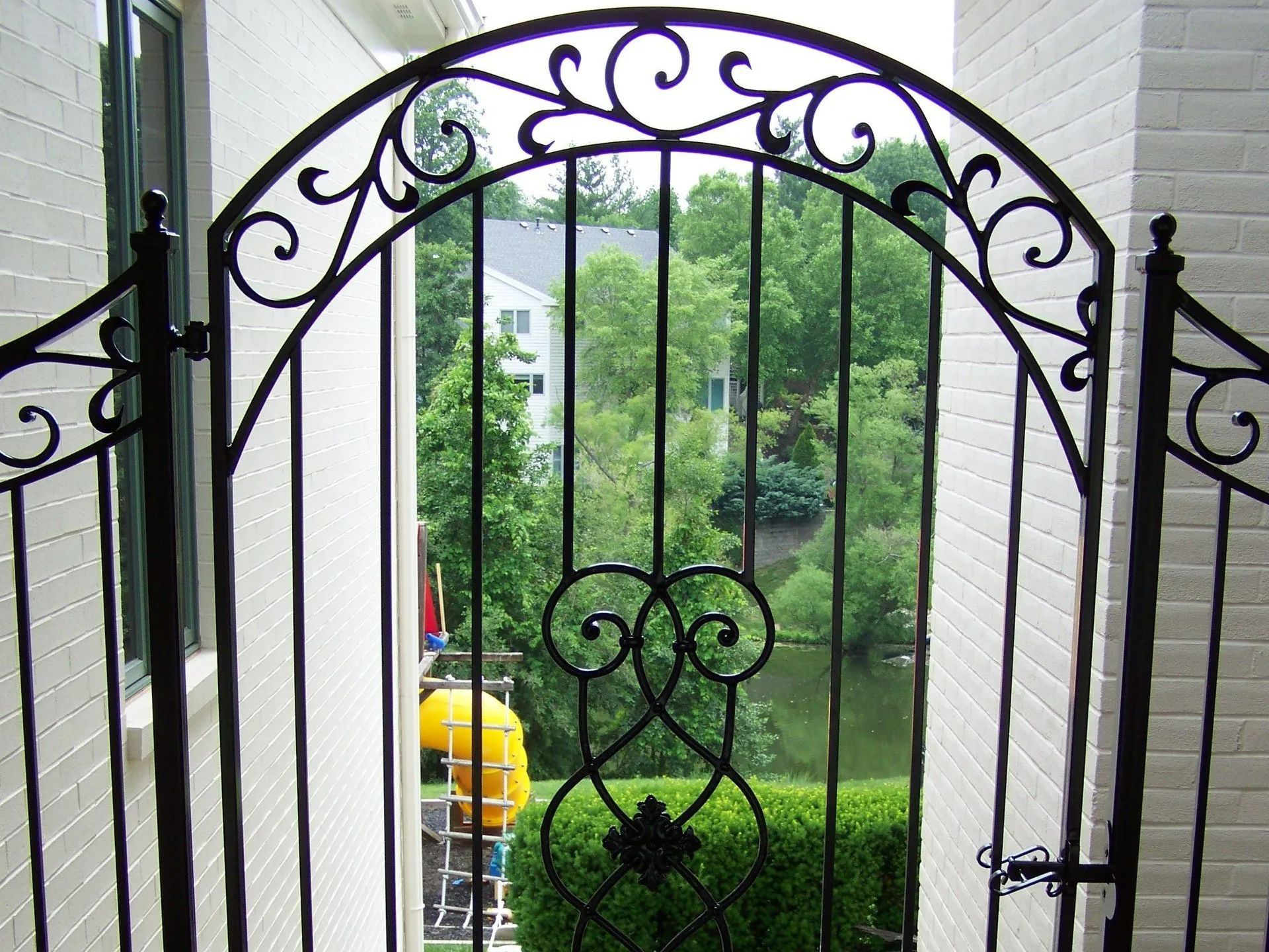 Black wrought iron gate with arched top, overlooking green trees and water.