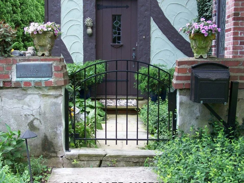 Wrought iron gate in front of a house entrance. Includes stone steps, mailbox, flower pots, and greenery.
