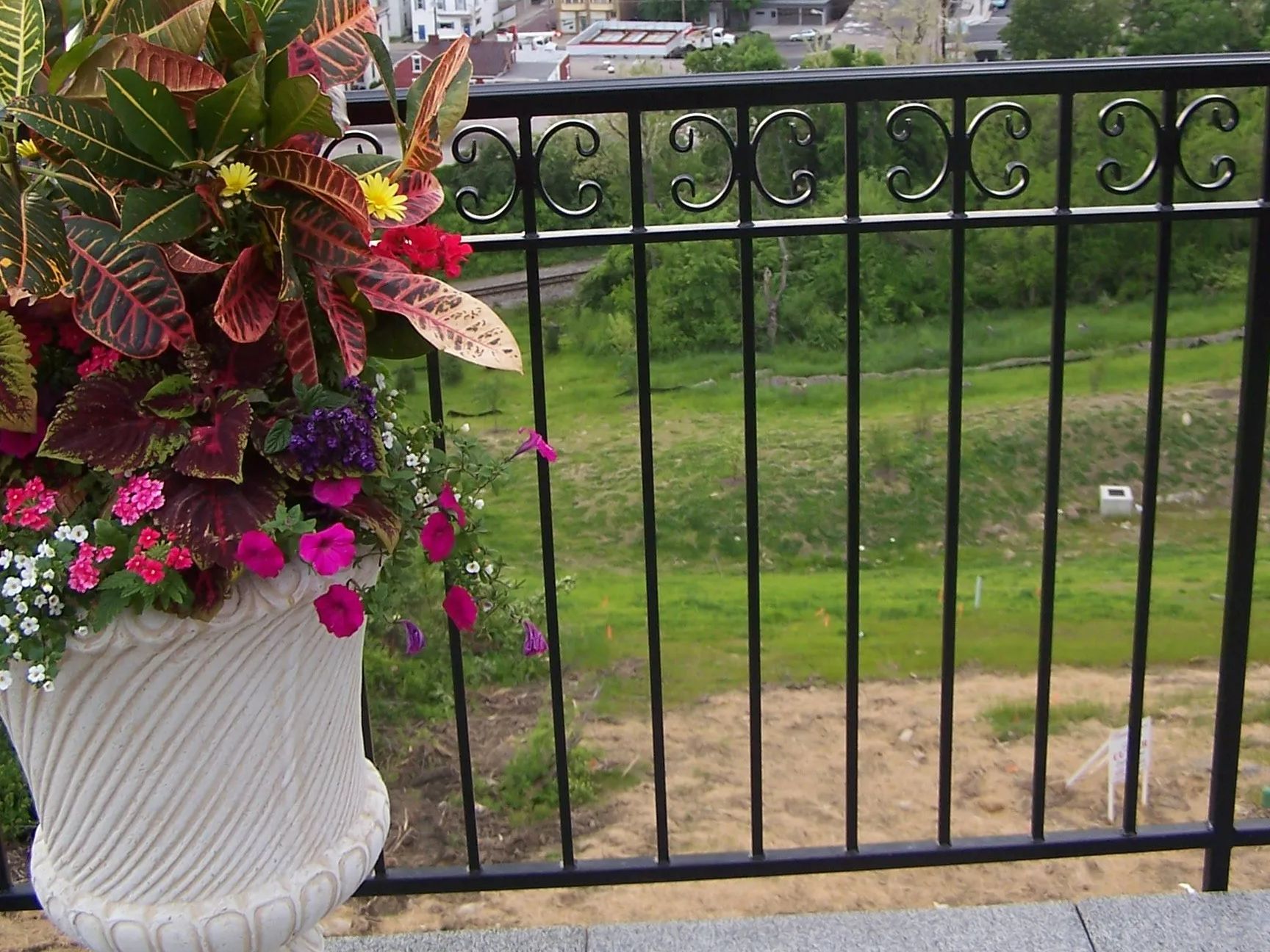 Potted flowers in a white urn on a patio with a black railing overlooking a green field and a distant cityscape.