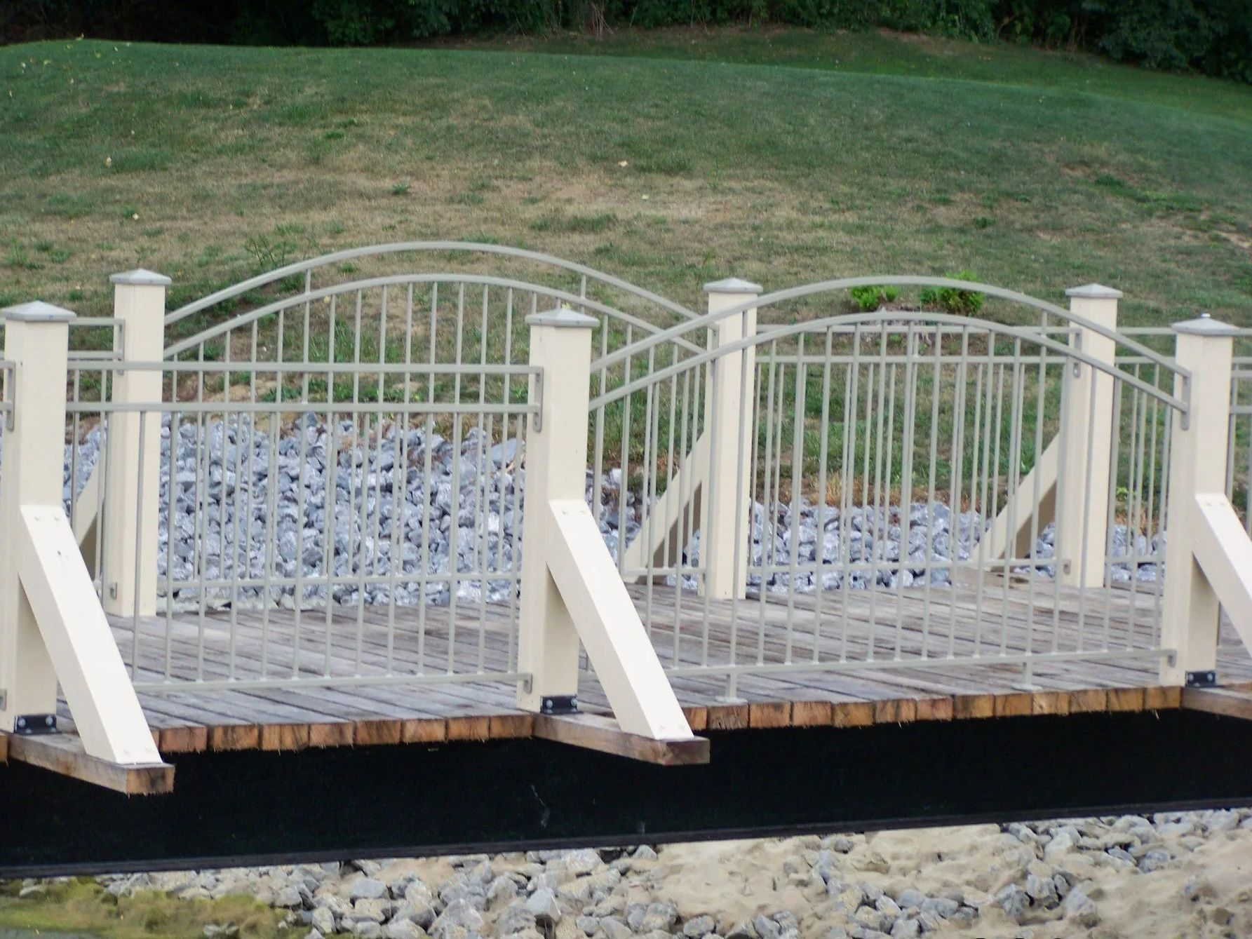 Cream-colored arched footbridge with metal railing over gray rocks and water, grassy bank in background.