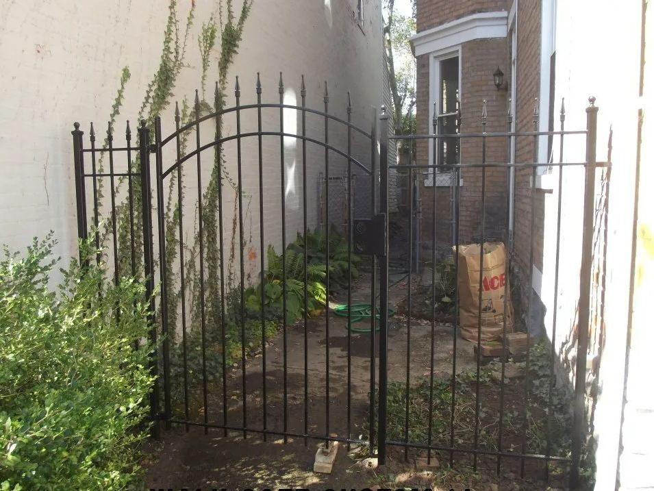 Black wrought iron gate between two buildings, partially enclosing a small garden.