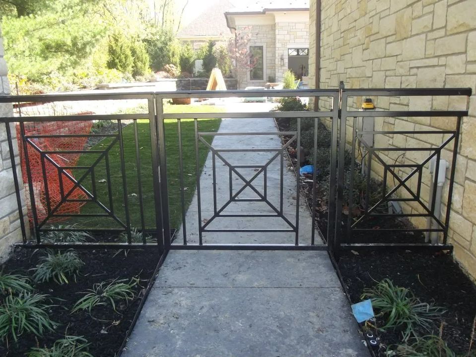 Black metal gate with geometric design, set between brick walls, leading to a yard with grass.