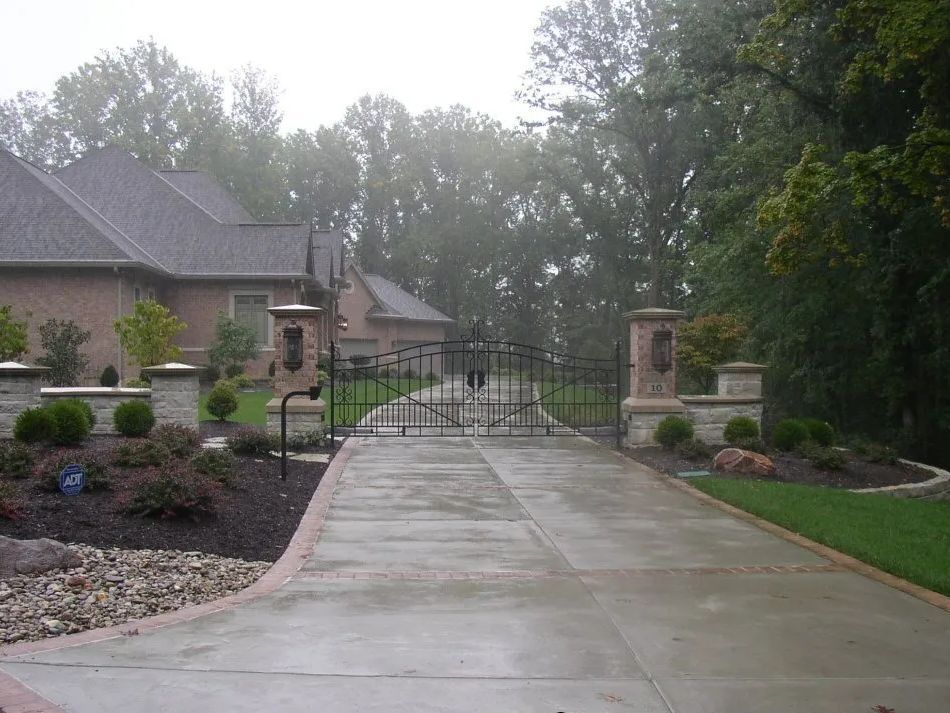 Gated driveway to a brick house on a rainy day. Concrete path, decorative columns, and landscaping.