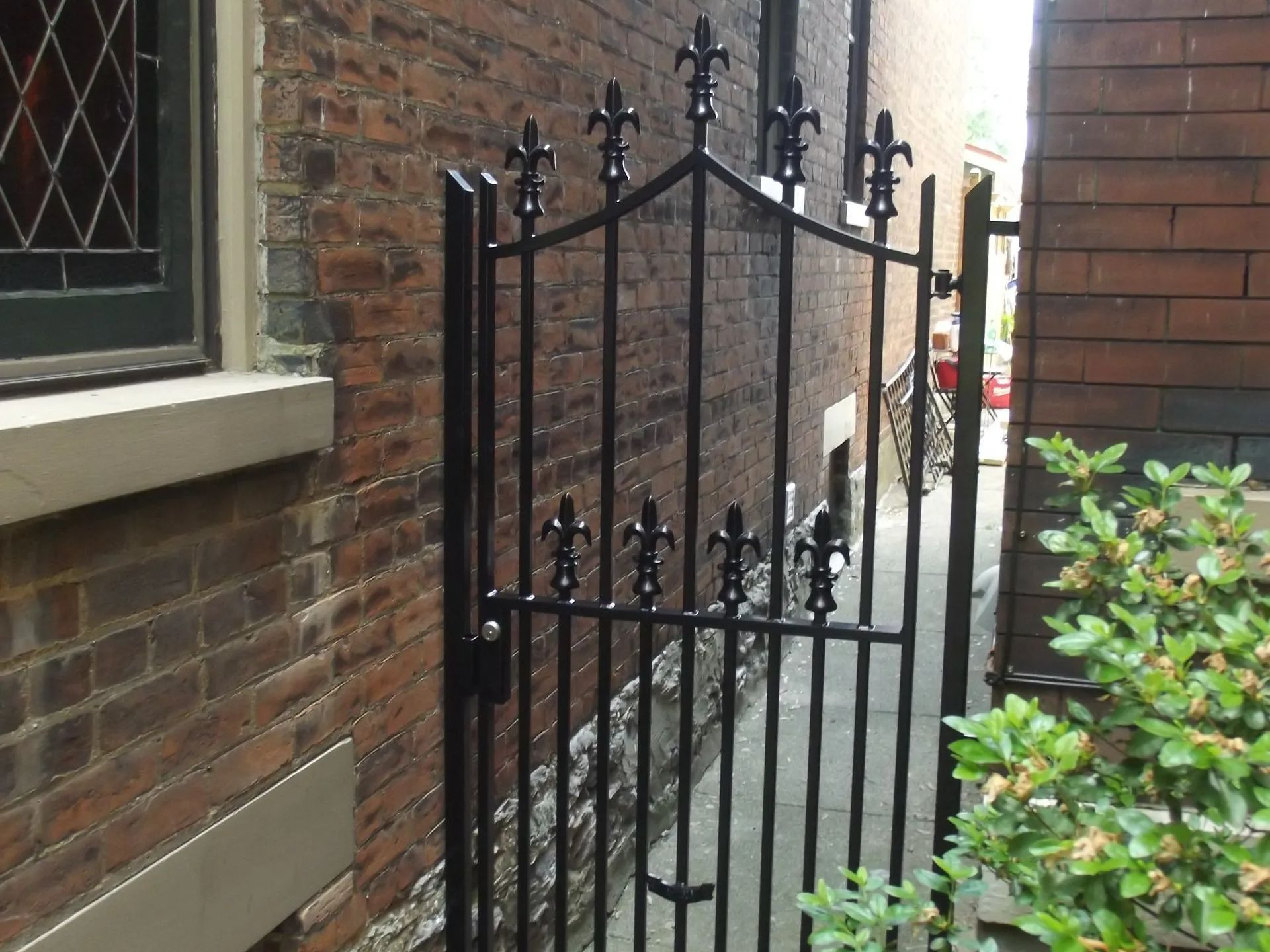 Black wrought-iron gate in a narrow brick alleyway. Fleur-de-lis detail on top.