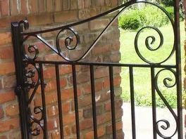 Black wrought iron gate with scroll designs against a brick wall and greenery.