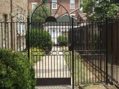 Black wrought-iron gate and fence lead to a backyard pathway; gate features an arched top, surrounding brick building.