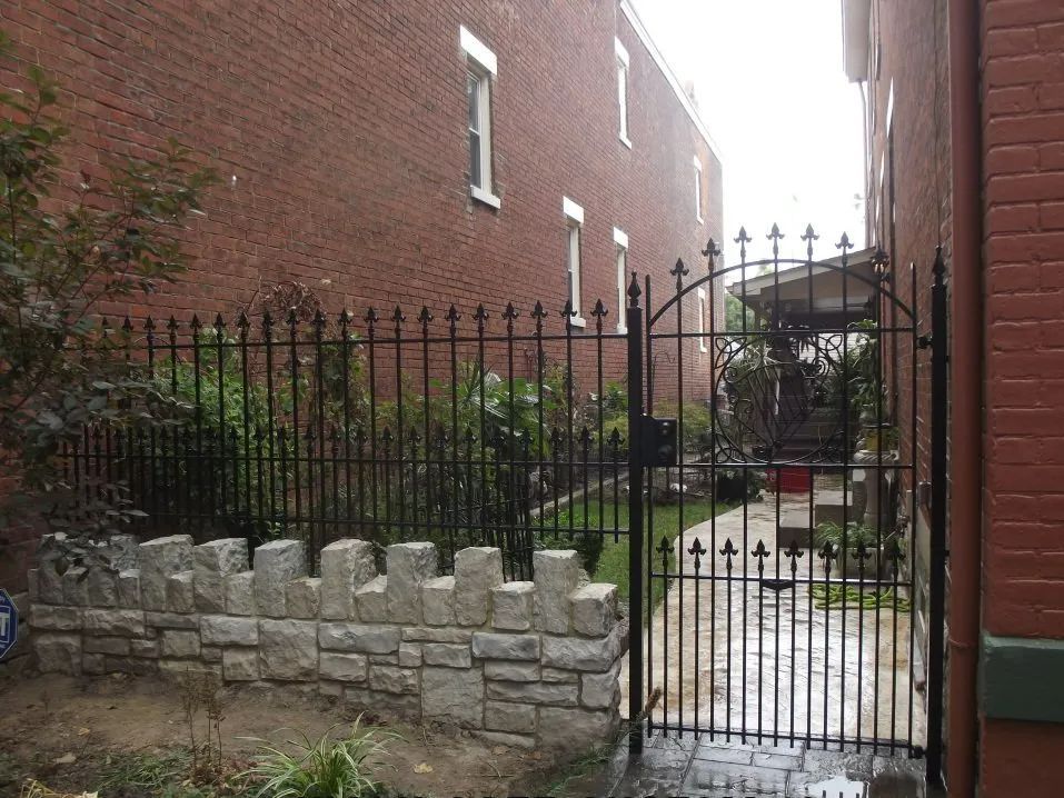 Black wrought-iron fence and gate lead to a walkway between brick buildings. A stone wall is in front.