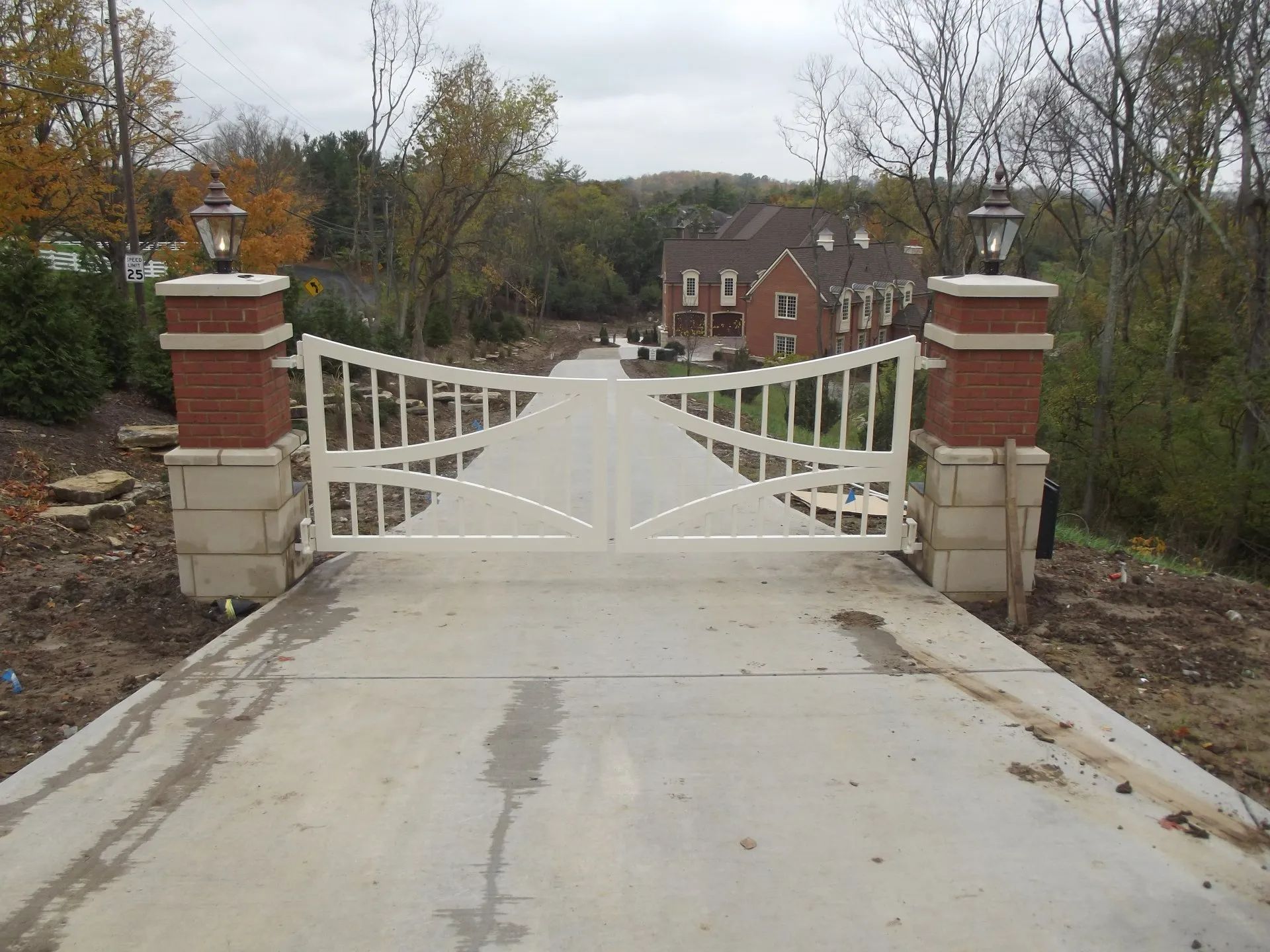 Driveway with white gate, brick and stone columns, leading to a brick house. Overcast sky.