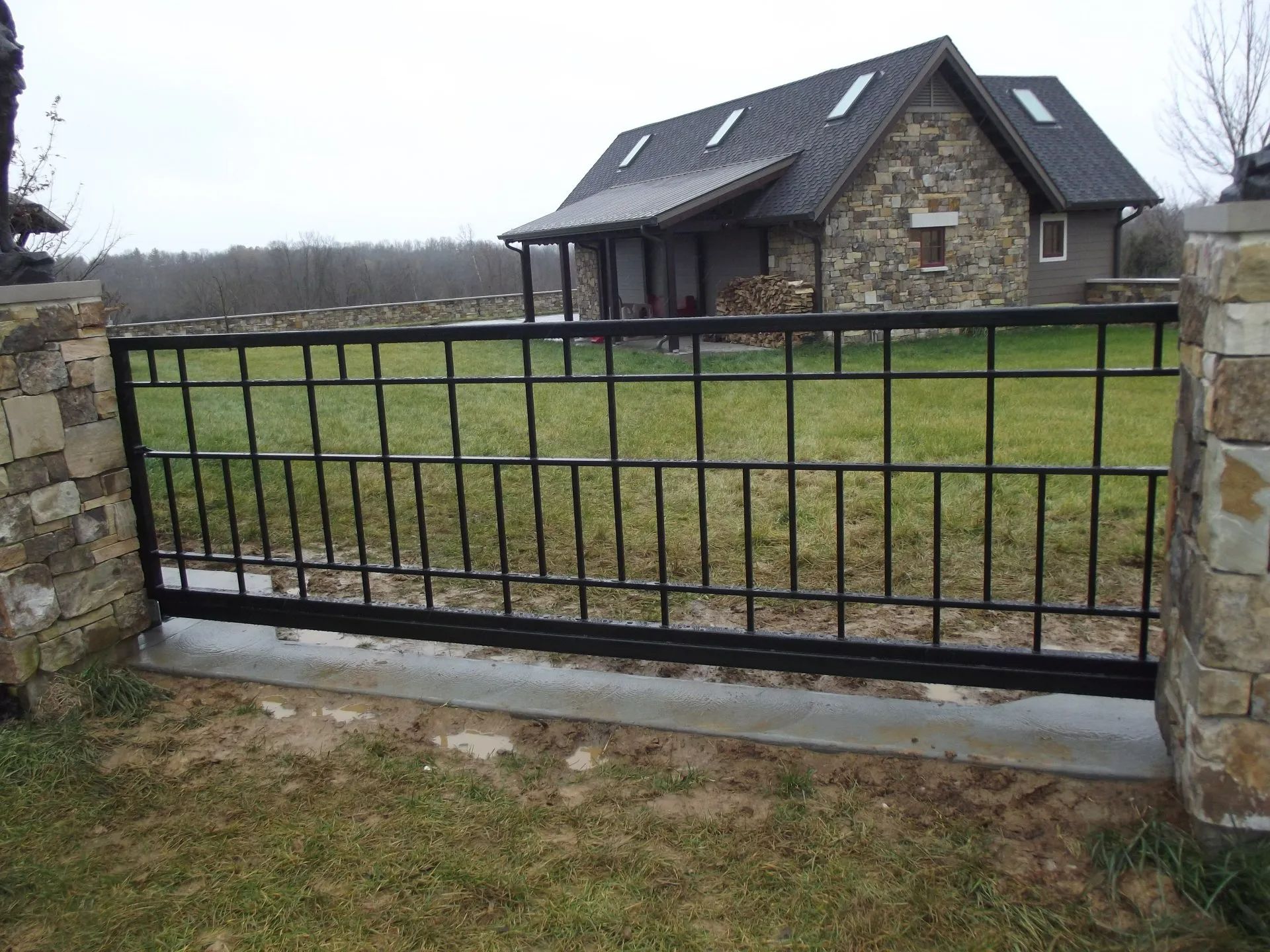 Black metal gate in front of a stone house with a dark roof.
