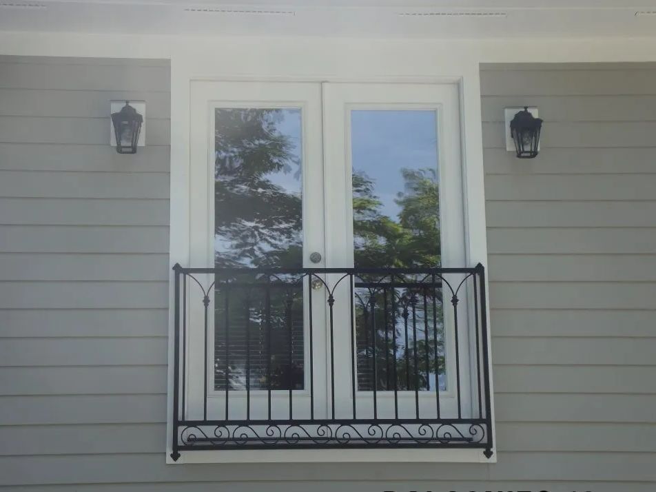 French doors with a wrought-iron balcony on a light gray siding building. Black sconces are mounted on the exterior.