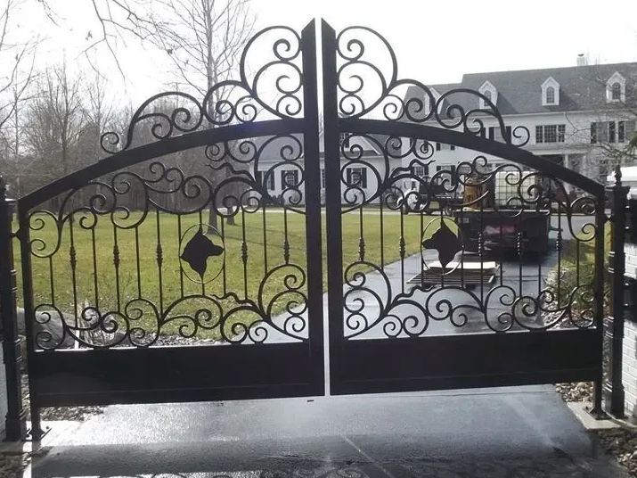 Ornate black wrought iron driveway gates with swirling scrollwork, leading to a green lawn and house.
