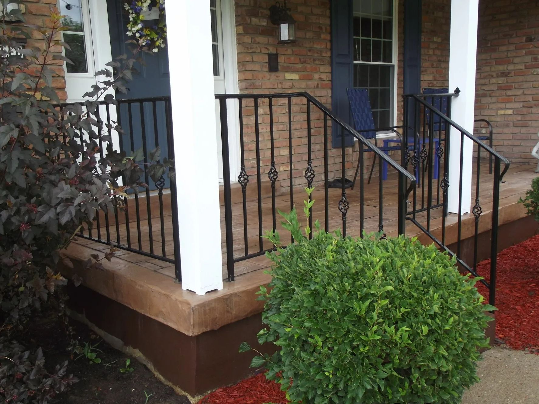 Black wrought iron railing on a brown and tan front porch with a small green bush.