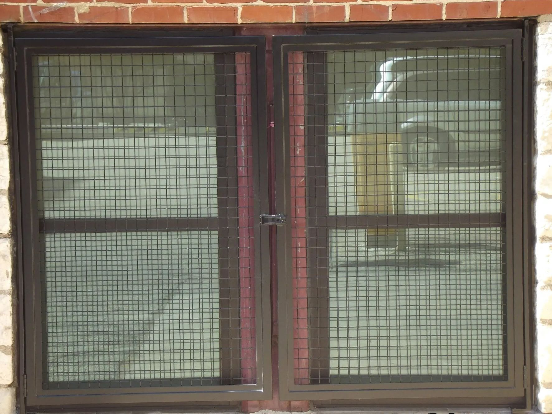 Brown-framed window with security grilles against a brick wall, reflecting a blurry outdoor scene.