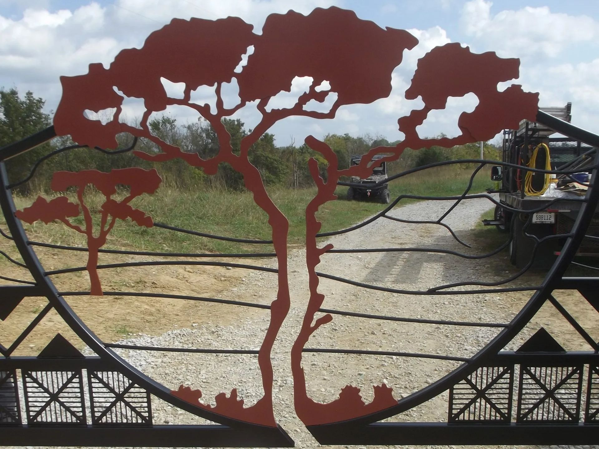 Decorative metal gate with a tree silhouette cutout and geometric designs, overlooking a gravel driveway and field.