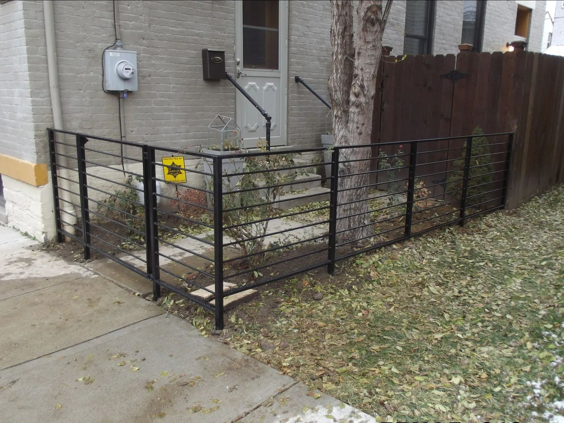 Black metal fence surrounds a small concrete porch with steps, a tree, and a brick building.