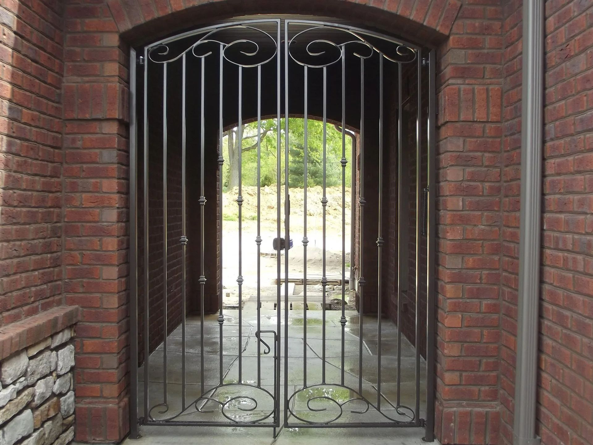 Wrought iron gates in a brick archway. Sunlight illuminates a path visible through the gates.