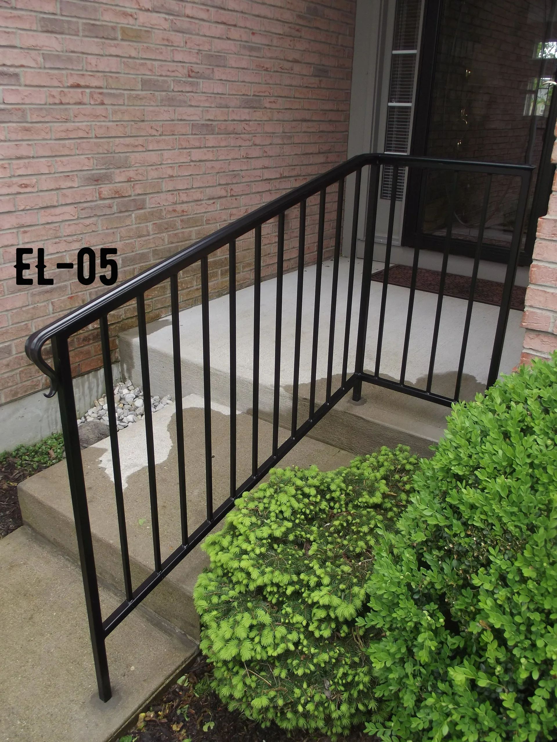 Black metal handrail on concrete steps leading to a door; brick wall and green bushes in view.