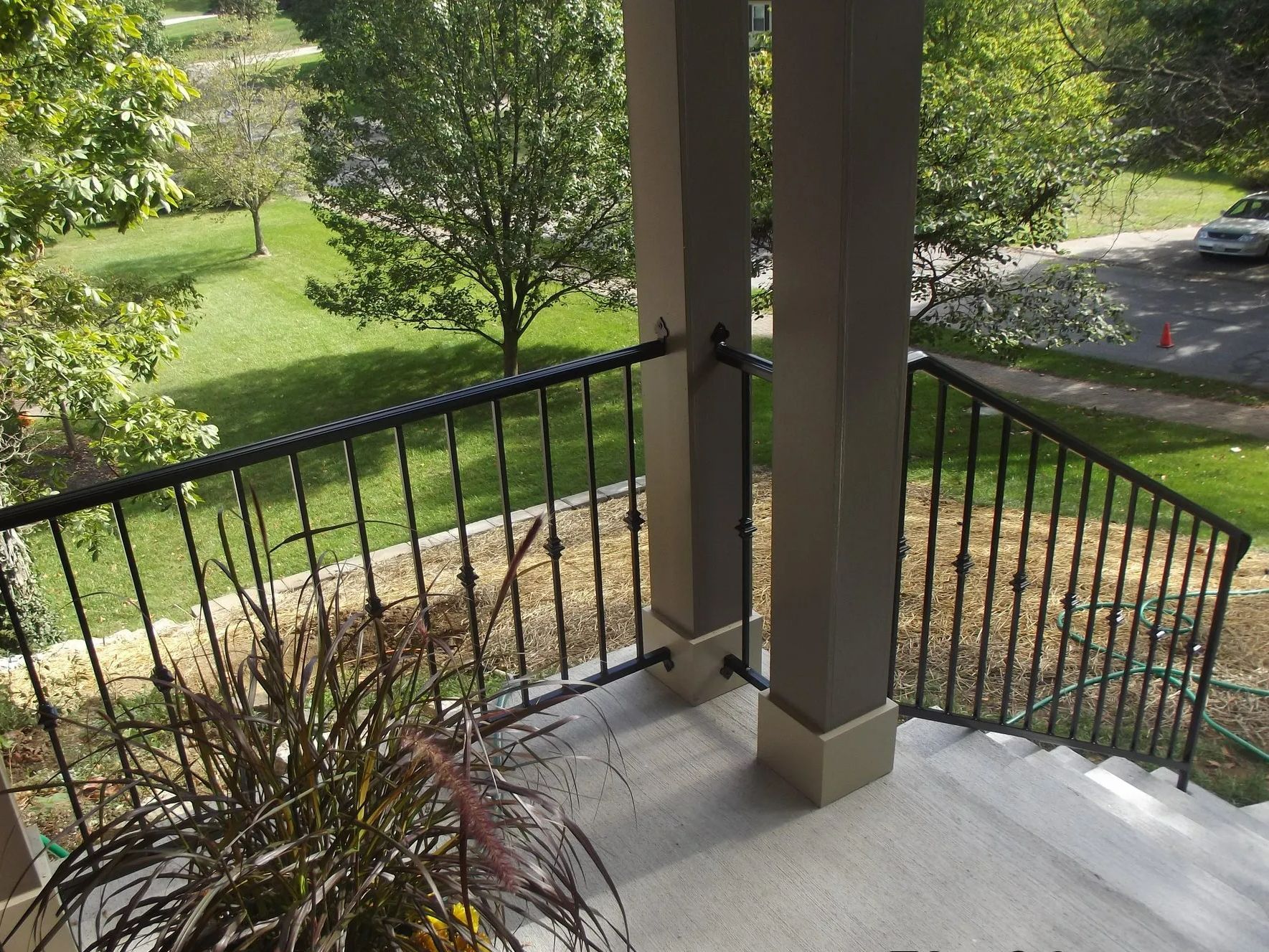 Black wrought-iron railing on porch, with brown support columns, overlooking a green lawn and trees.
