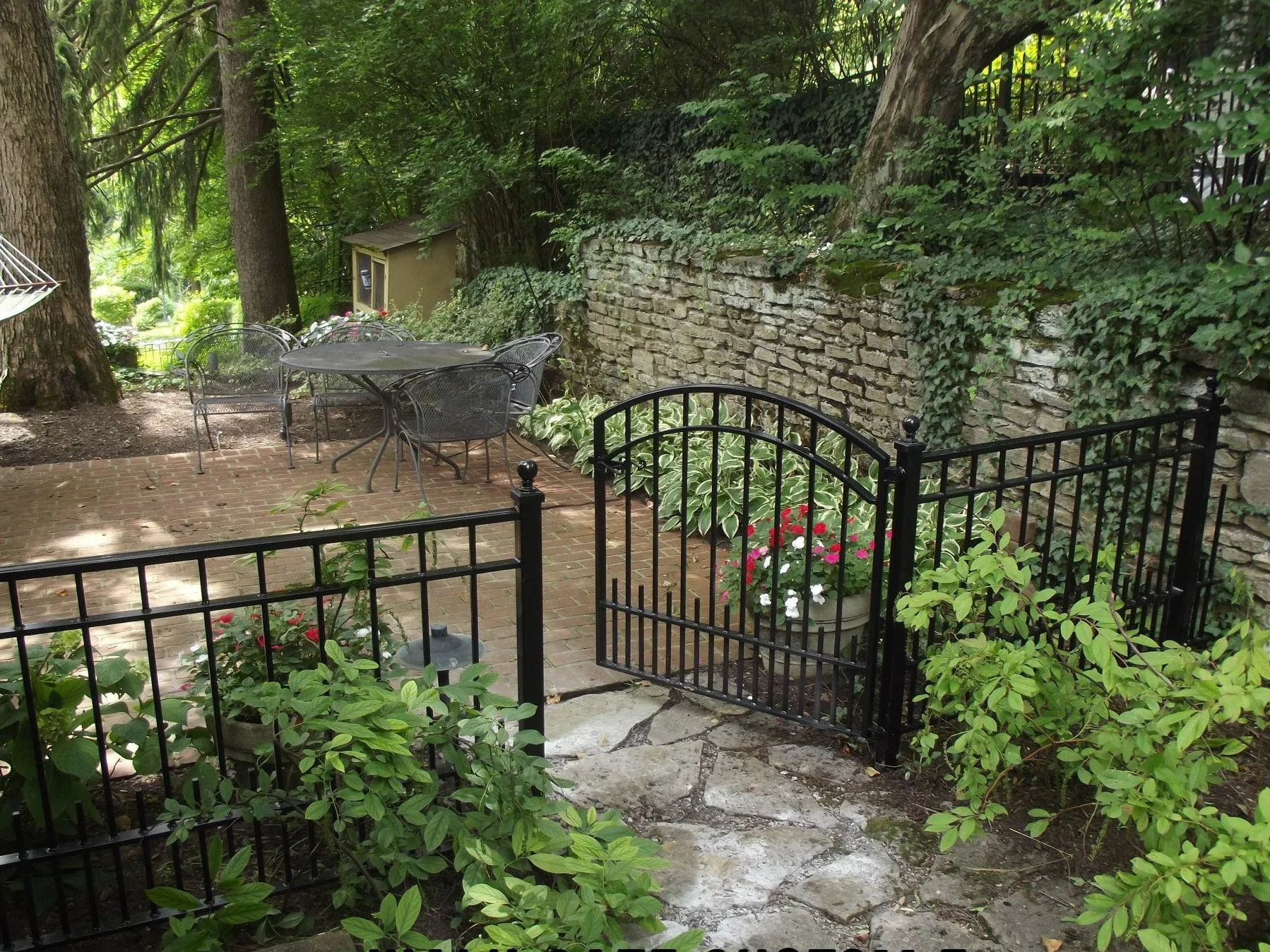 Black wrought iron fence with gate leading to a garden with stone wall, trees, and patio furniture.