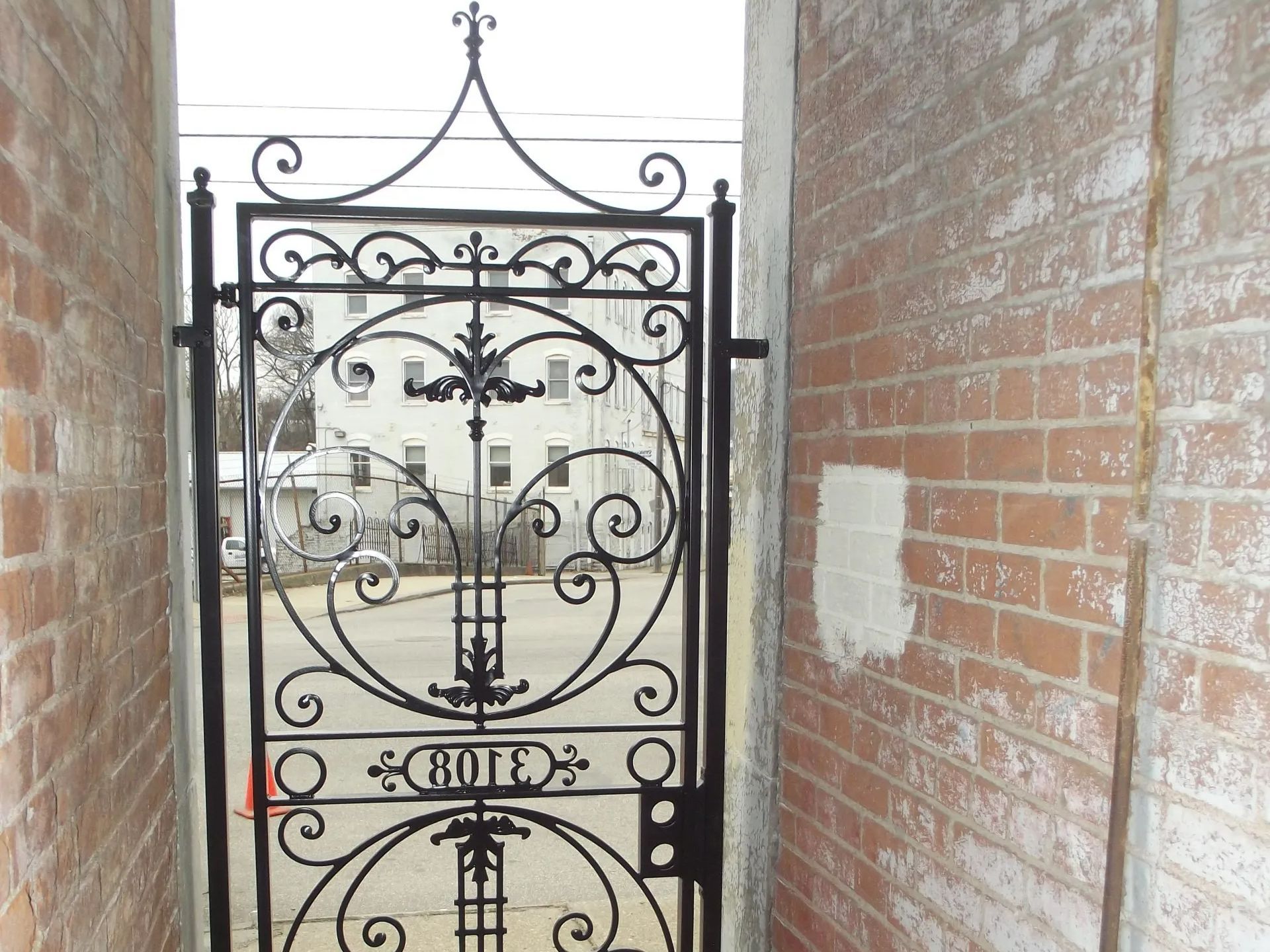 Black ornate gate between brick walls, with a street and building visible through the gate.