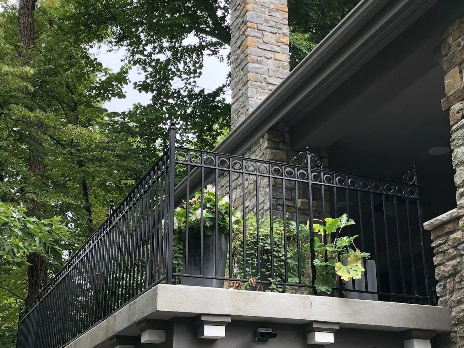 Black wrought-iron railing on a concrete balcony with potted plants, next to a stone chimney and overhanging roof.