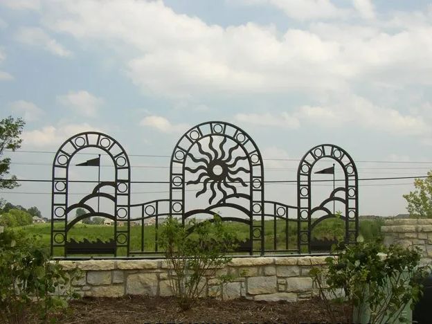 Black metal fence with sun design atop a stone wall, in front of greenery and a cloudy sky.