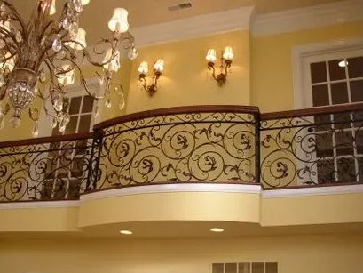 Balcony with ornate wrought iron railing, chandelier, and wall sconces in a yellow-painted interior.