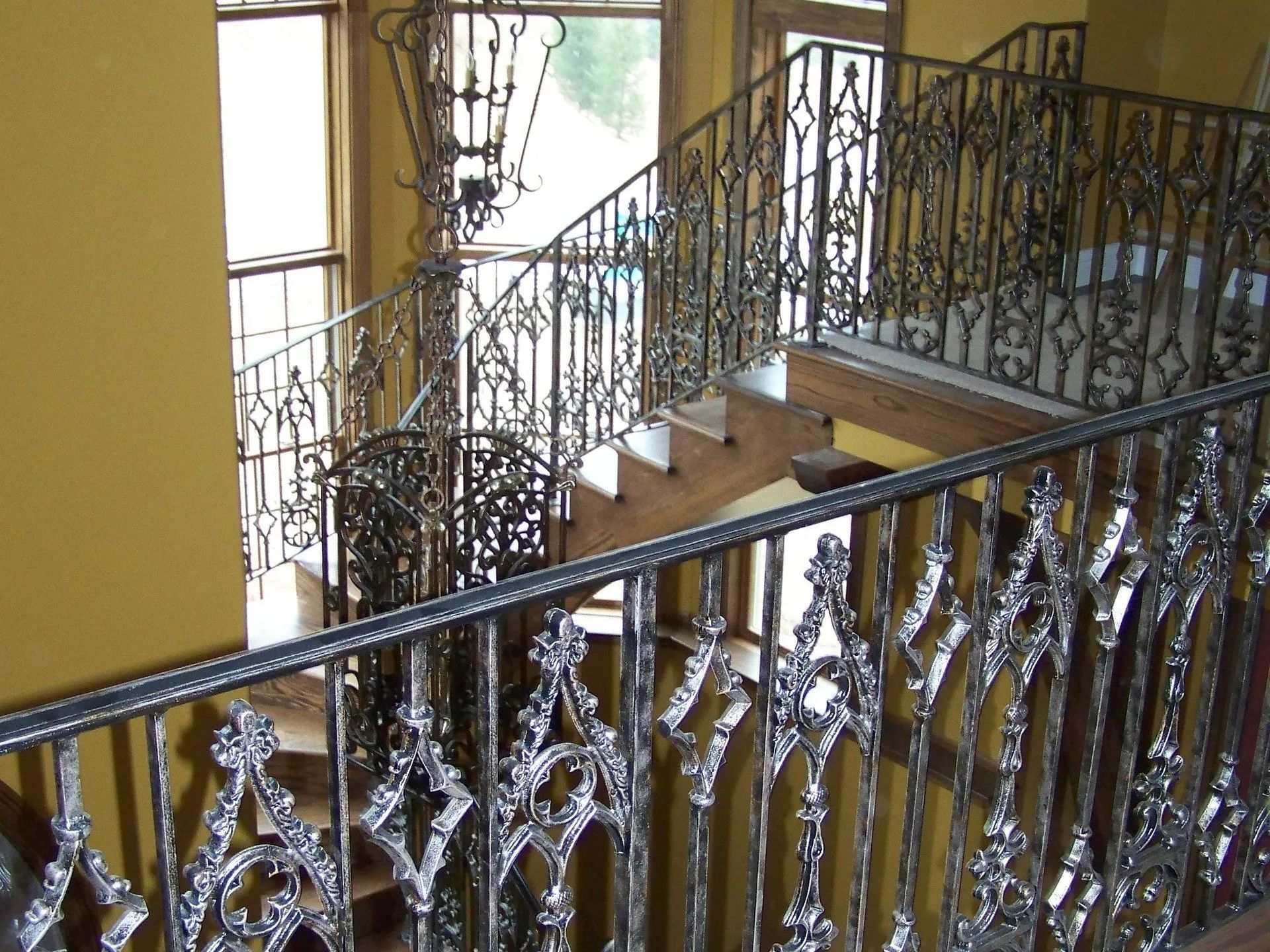 Wrought iron staircase with ornate railing, steps leading up. Yellow walls and natural light from windows.