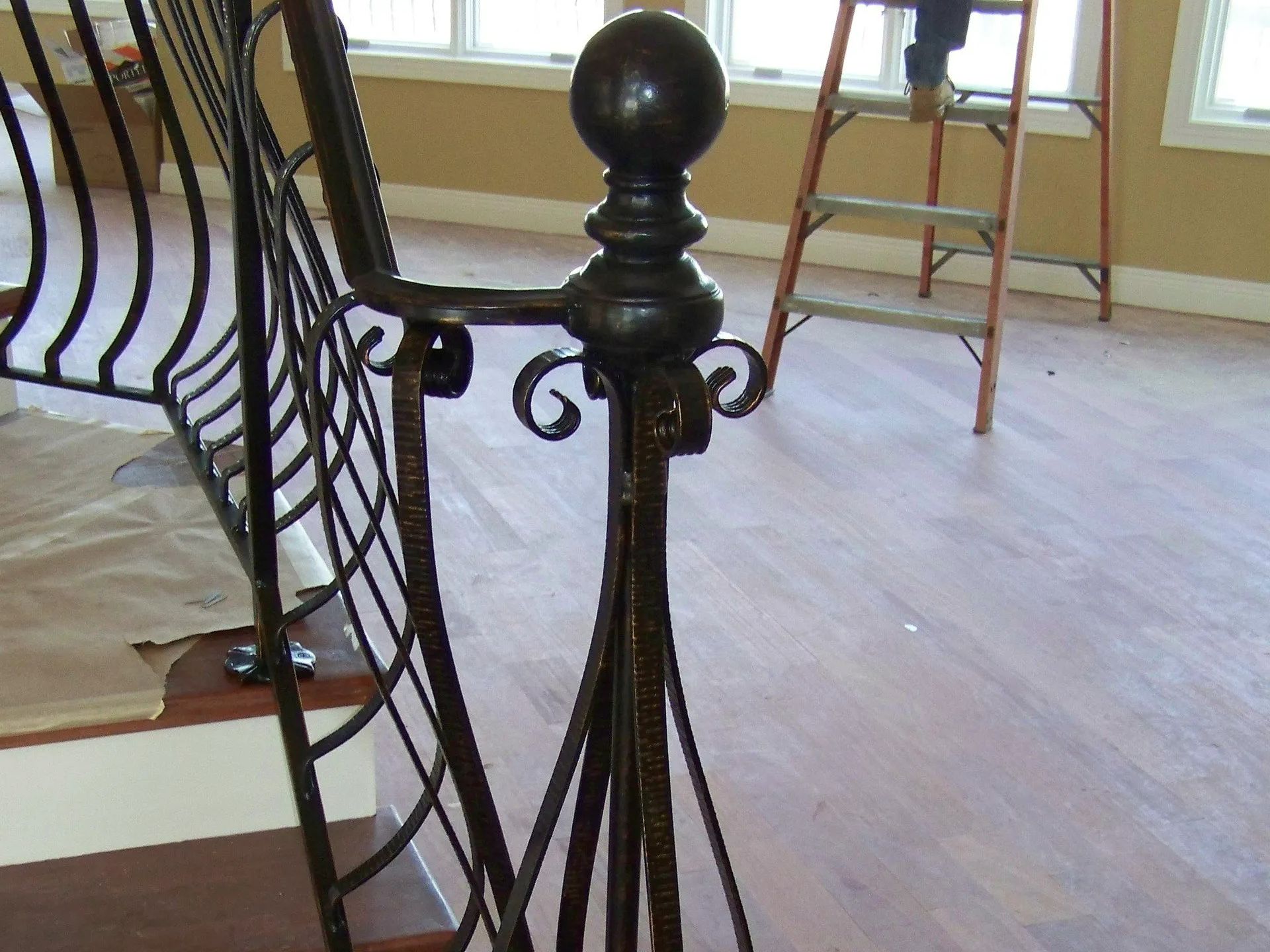 Ornate wrought-iron stair railing with a decorative finial. Interior setting with wooden floor and ladder in the background.