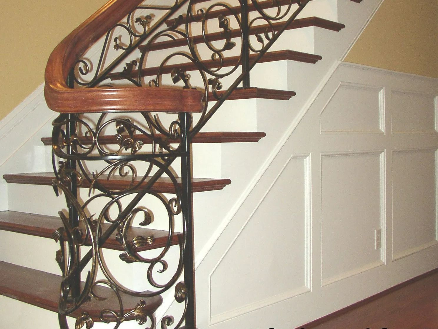 Staircase with wooden steps and railing, ornate black iron balusters, and white paneled wall.