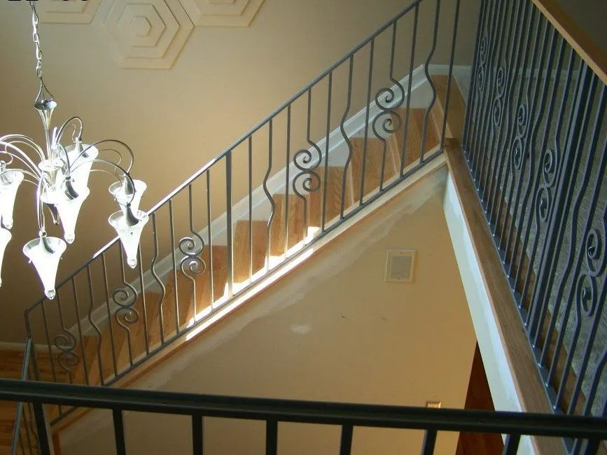 Iron railing on a staircase with decorative scrolls, light-colored walls and ceiling, chandelier hanging above.