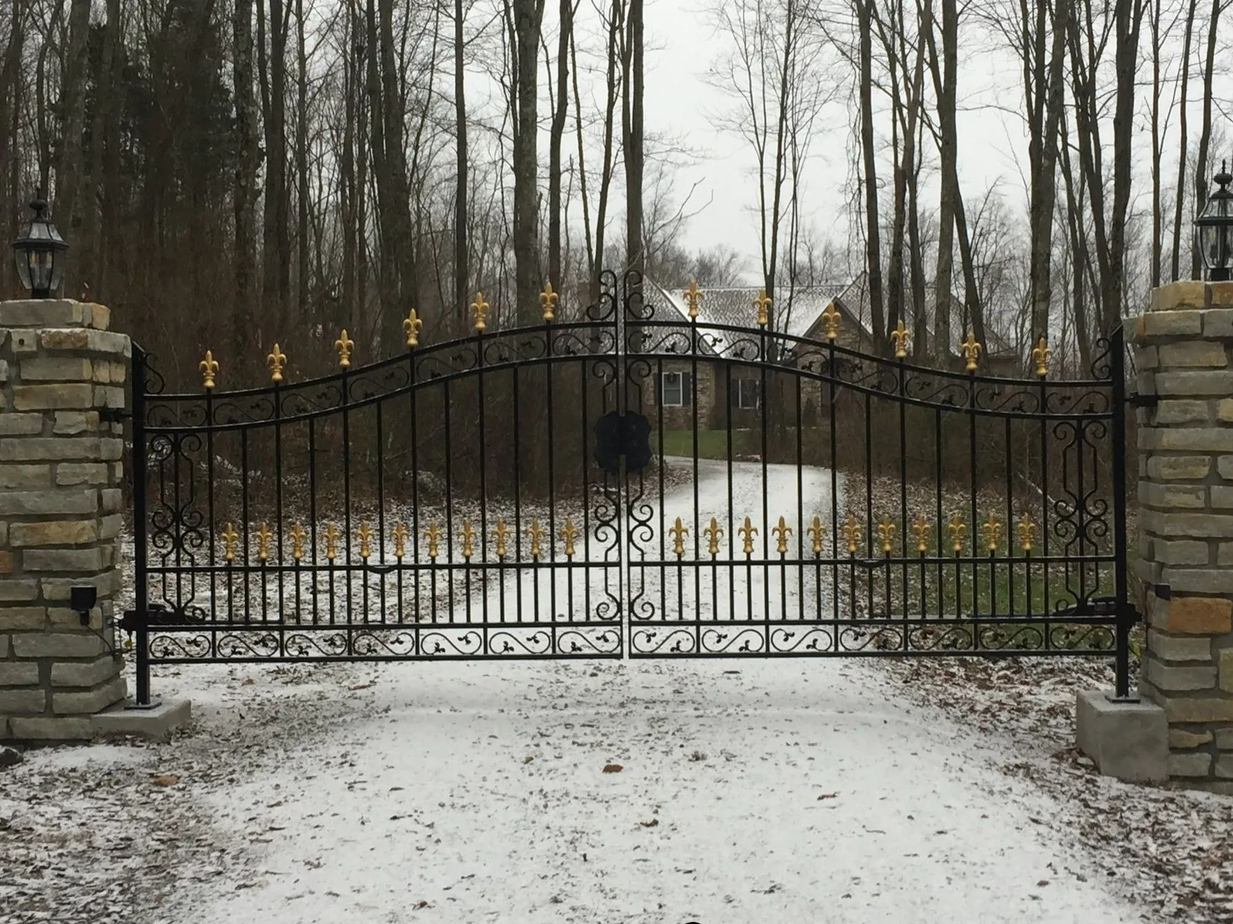 Black wrought iron gate with gold accents, set between stone pillars, leads to a snow-covered driveway.