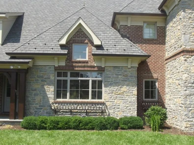 Stone and brick house exterior with a gray roof and manicured bushes.