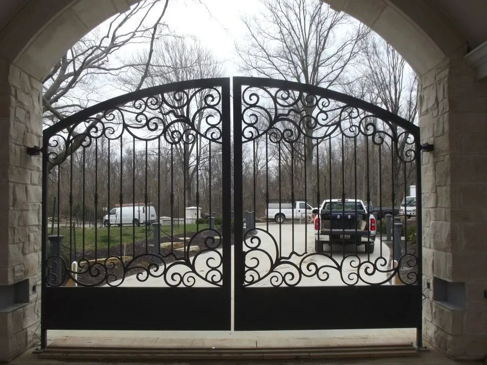 Ornate black wrought iron gates, arched at top, open to a driveway with vehicles, set in stone entryway.