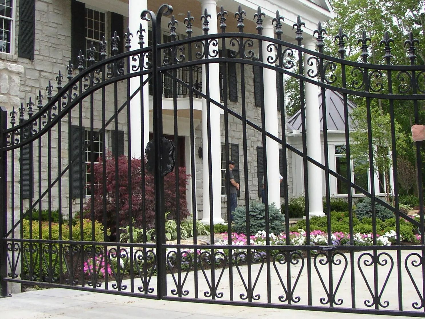 Black wrought iron gate in front of a large, two-story house with columns and landscaping.
