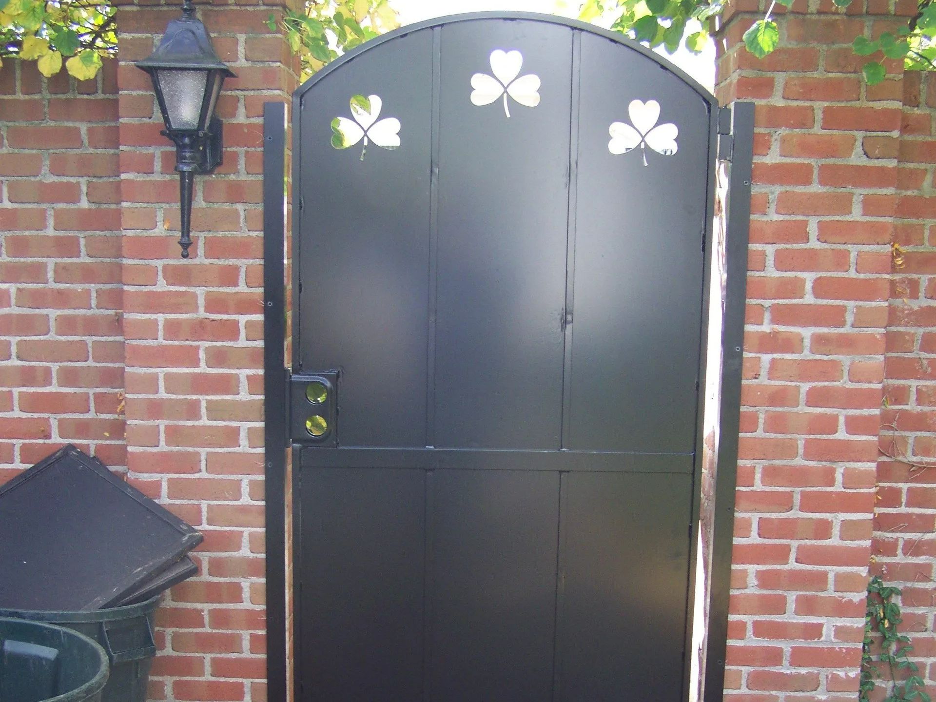 Black metal gate with clover cutouts set in a brick wall, with a black lamp to the left.