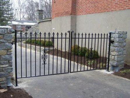 Black metal driveway gate between stone pillars, set in concrete, with brick building backdrop.