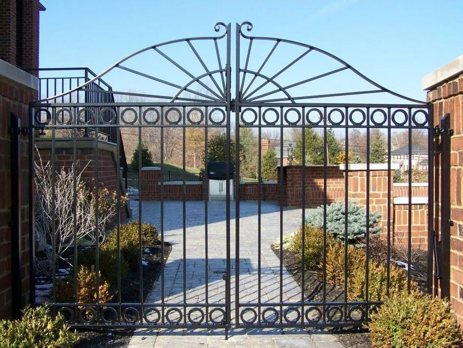 Black iron gate with arched top, circles, and vertical bars between brick columns in front of a house.