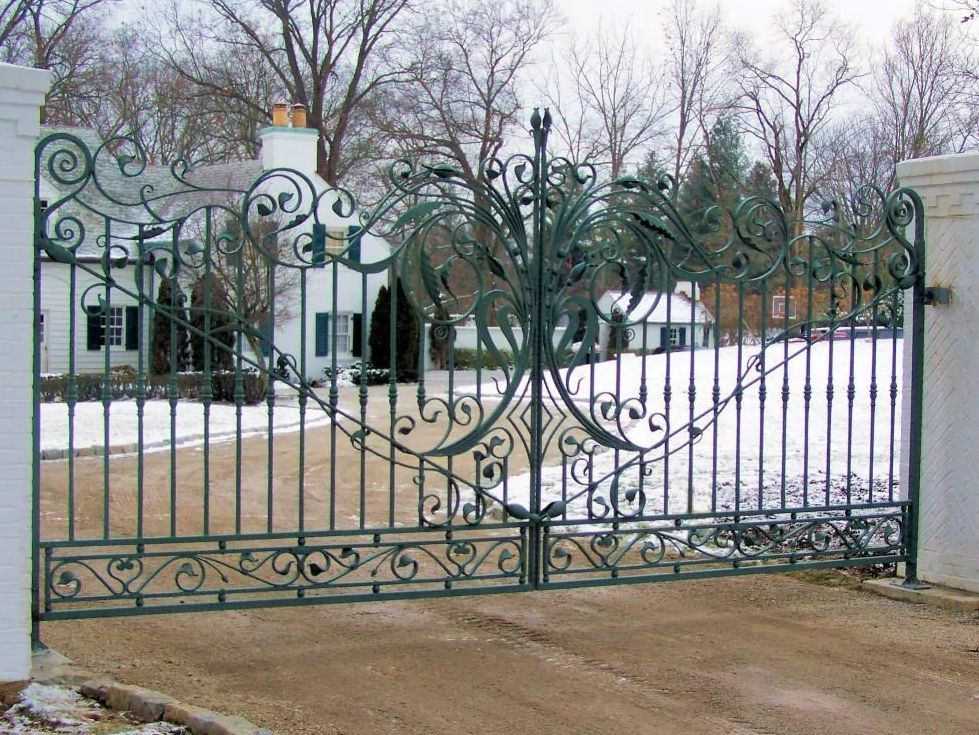 Ornate green wrought iron driveway gates with a white house in the background on a snowy day.