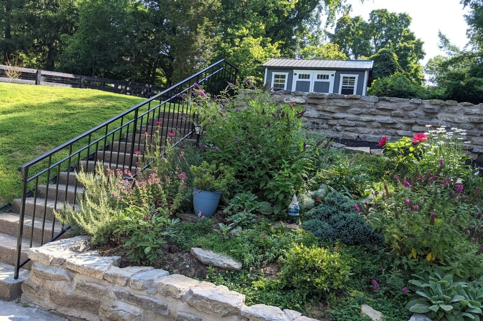 Stone steps with black railing leading up a grassy hillside garden with a small gray building in the background.