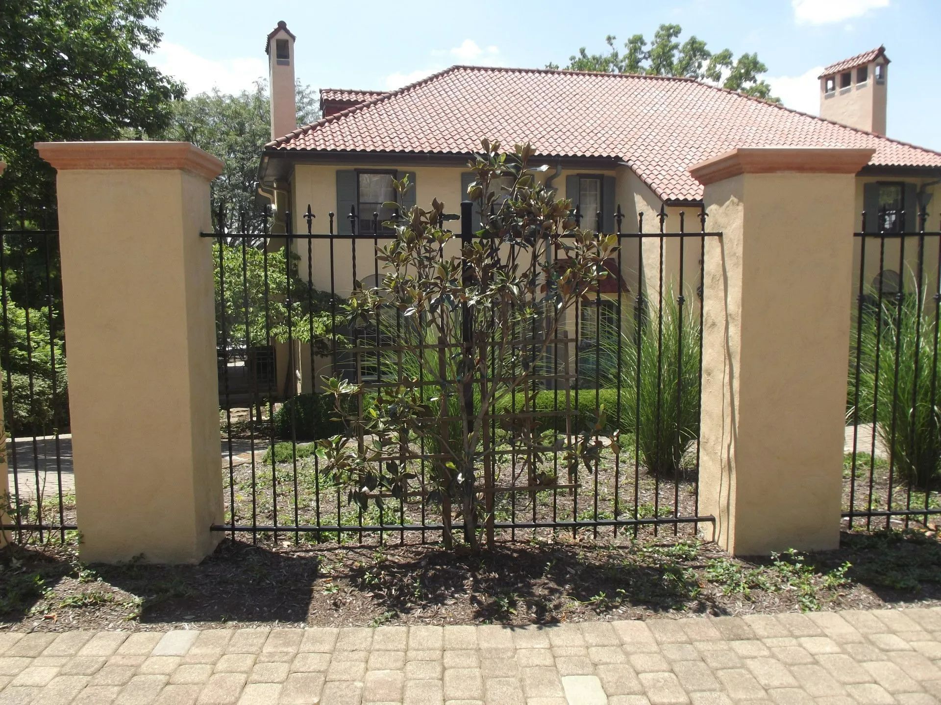 Tan stucco pillars with brown caps frame a black iron fence. A house with a red tile roof is in the background.