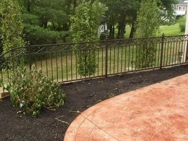 Black metal fence bordering a red-orange patio, with trees and lawn in background.